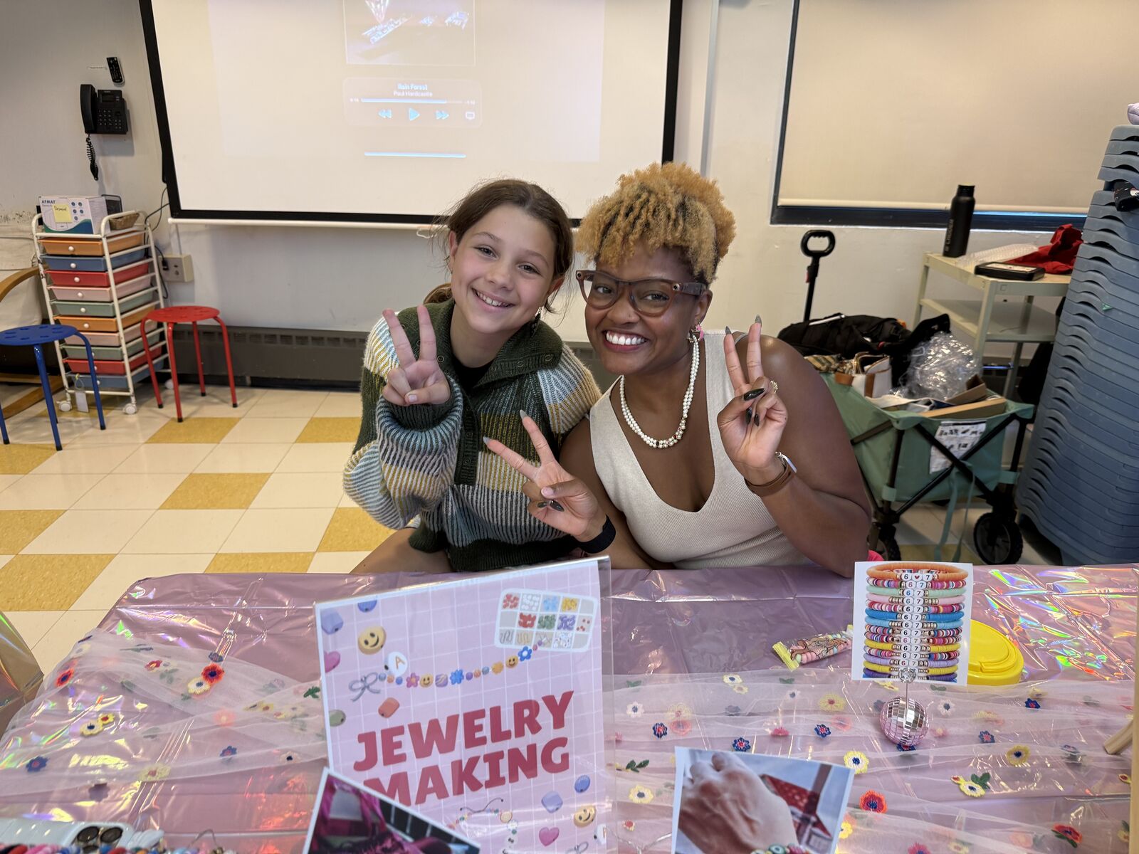 Gabriell and a smiling student throwing peace signs at a jewelry making table